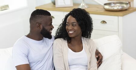Black millennial couple smiling and looking at each other, sitting on sofa at home, panorama