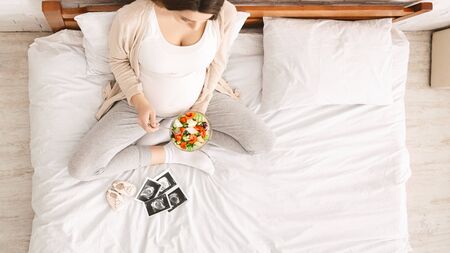 Healthy snack . Young pregnant woman eating fresh vegetable salad, looking ultrasound photos of baby...