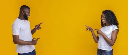 Happy african american couple pointing fingers at free space in the middle, yellow studio background