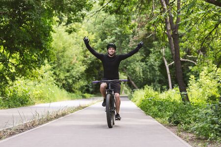 Success concept. Professional male cyclist celebrating victory, raising hands up while riding by finish trail in park, empty spaceの写真素材
