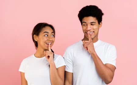 Amazed teen couple making silence sign, keep forefingers on lips over pink background
