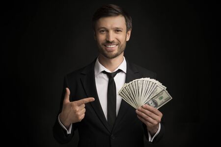Income. Smiling Businessman Pointing Finger At Money Cash In His Hand Standing Over Black Studio Background.の写真素材