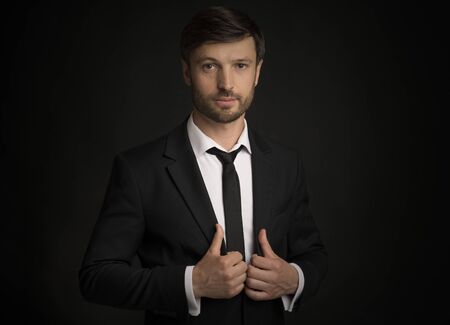 Confident Businessman Looking At Camera Holding Jacket Lapels Posing Over Black Background. Studio...