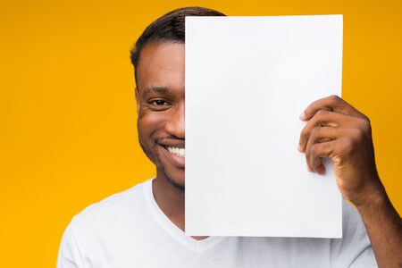 Smiling African American Man Holding Blank Paper Sheet Covering Half Of His Face Standing Over Yellow Background. Mockupの写真素材