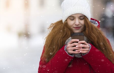 Girl with closed eyes holding hot drink, enjoying winter day under the snowfall, copy space
