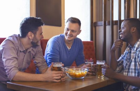 Three young multiracial men having good time at bar, talking and drinking beerの写真素材