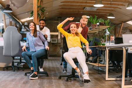 Time for break. Joyful colleagues having fun at workplace, making office chair race in coworking...