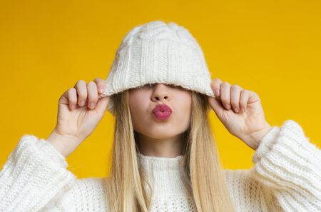 Playful blonde woman pulling down woolen white hat, showing kissing lips, yellowの写真素材