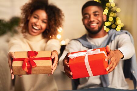 Excited afro couple holding xmas gift boxes and stretching them to camera in front of Christmas treeの写真素材