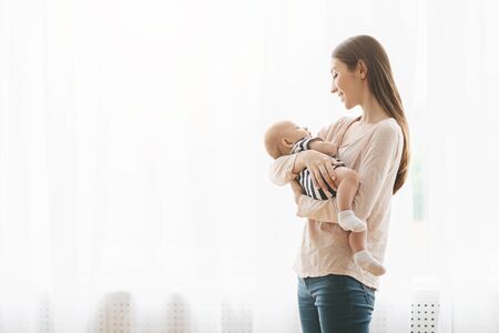 Young mother holding her newborn child, lulling baby while standing near window at home, free spaceの写真素材