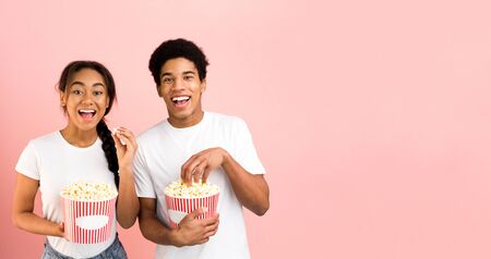 Overjoyed teen couple enjoying movie with buckets of popcorn, pink background with free spaceの写真素材