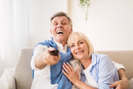 Watch tv. Excited mature couple switching channels with remote controller, resting at home