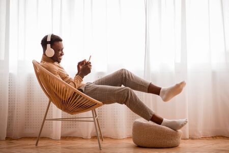 Weekend At Home. Happy Afro Guy In Headphones Using Smartphone Sitting On Modern Chair Near Window...