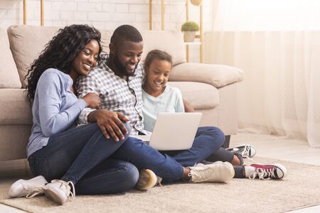 Planning vacation together. African father, mother and daughter using laptop at home, sitting on floor, copy spaceの写真素材
