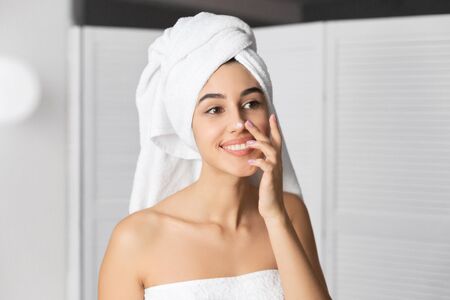 Cheerful Girl With Towel On Head Applying Face Cream On Nose Moisturizing Skin Standing In Bathroom. Selective Focusの写真素材