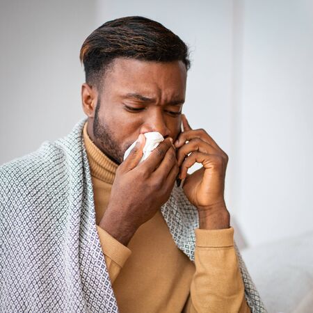 Cold And Flu. African American Man Blowing Runny Nose In Paper Tissue Talking On Phone Wrapped In Blanket At Home.の写真素材