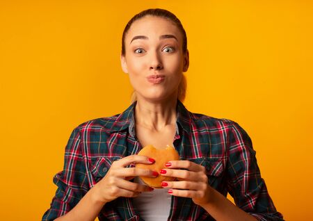 Slim Girl Eating Burger Having Cheat Meal Day Standing On Yellow Studio Background. Unhealthy Diet