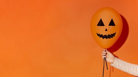 Halloween party. Balloon with drawing face in woman hand on orange background, panorama, copy spaceの写真素材