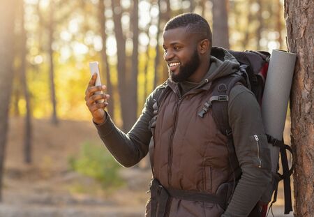 Cheerful black guy with camping backpack taking photos of autumn forest on smartphone, empty space