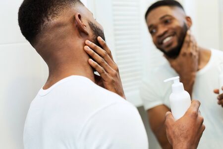 Facial care. Young black man applying moisturizer on beard in bathroom, looking in mirrorの写真素材