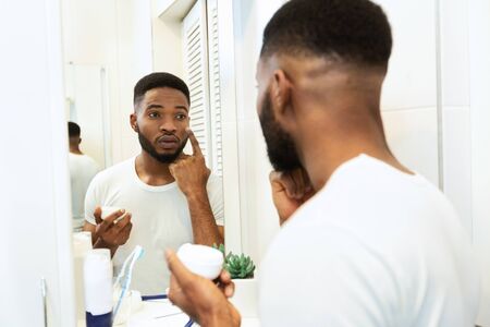 Male skin care. Handsome african man applying moisturizing cream in bathroomの写真素材