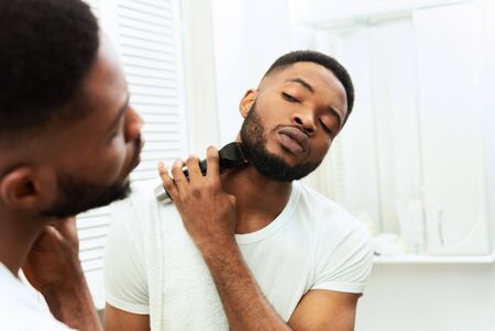 Grooming concept. Young black man looking at mirror and shaving beard with trimmer or electric...