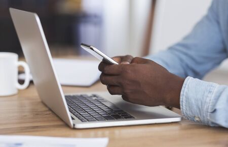 Multitasking concept. Unrecognizable african american employee typing message on smartphone and...