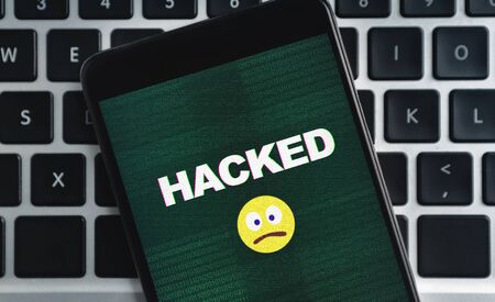 SIM-swap attack concept. Smartphone with hacked inscription and sad emoji icon on screen over laptop...