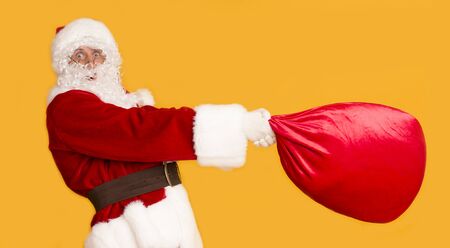 Funny Santa trying to pick up big heavy bag full of presents on orange background, panoramaの写真素材