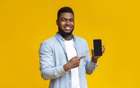 Handsome african american guy demonstrating smartphone with black screen and pointing on it, yellow background with free spaceの写真素材