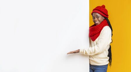 Empty Space For Advertising. Black cheerful girl in red knitted set directing with palm to empty white board, yellow background, panoramaの写真素材
