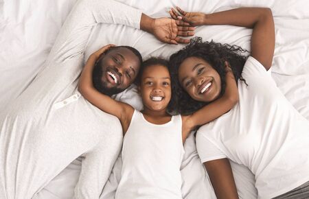 Family weekend. Smiling black mother, father and daughter lying on bed together, relaxing at home.の写真素材