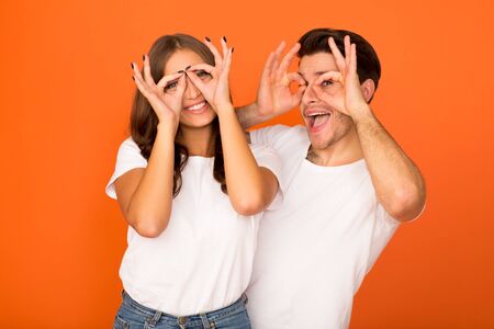 Crazy couple making funny glasses, holding fingers near eyes, orange studio background