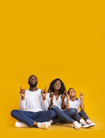 Happy black family of three sitting on floor and pointing upwards at copy space over yellow backgroundの写真素材