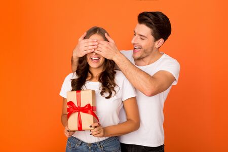 Loving man making surprise for girlfriend, closing her eyes over orange backgroundの写真素材