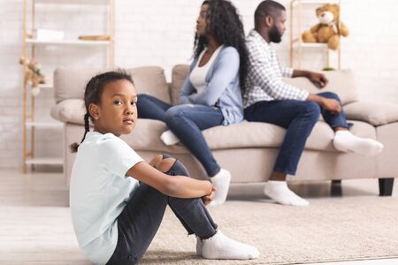Family problems concept. Upset little black girl sitting separately from parents after their arguing, feeling depressed and frustrated.の写真素材