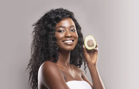 Beauty Treatments for Skin and Hair Concept. Portrait of attractive black woman holding avocado half...