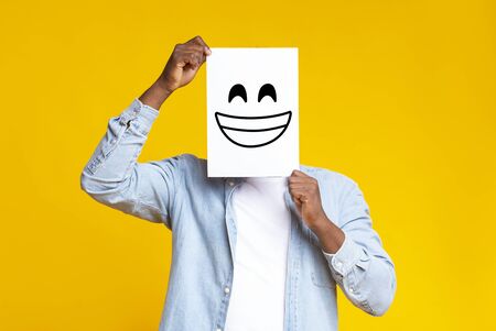 Happiness and success concept. Black male hiding face behind sheet of paper with drawn happy emoticon over yellow background in studio, copy spaceの写真素材