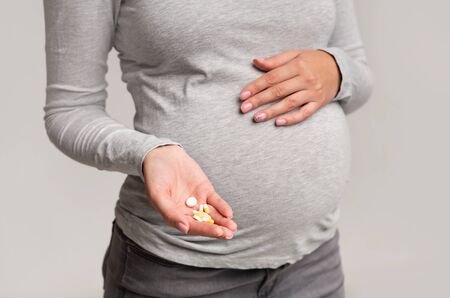 Unrecognizable Pregnant Girl Showing Pills In Hand Touching Belly Standing Over Gray Studio Background. Croppedの写真素材