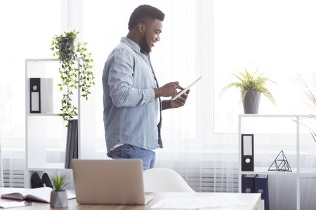 Successful african american entrepreneur holding digital tablet, browsing internet against window in modern office, side viewの写真素材