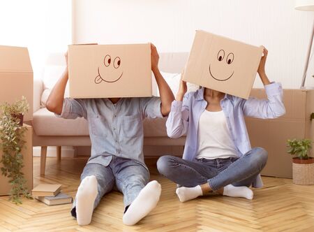 Moving House. Unrecognizable Couple With Cardboard Boxes With Smiley Faces On Head Sitting On Floor...