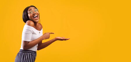 Excited African American Girl Pointing Finger At Open Palm Standing Over Yellow Background. Studio Shot, Panorama With Empty Spaceの写真素材