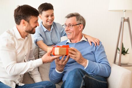 Mature Father And Son Congratulating Grandpa Giving Him Birthday Gift Sitting On Sofa At Home. Selective Focusの写真素材