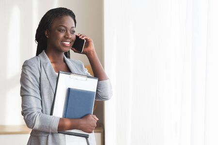 After successful interview. Black female job seeker talking on cellphone and holding folder with CV in her arms, standing near window in officeの写真素材