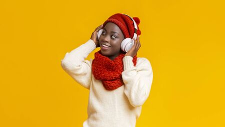 Excited afro winter girl with wireless headset looking aside, yellow studio background, copy spaceの写真素材