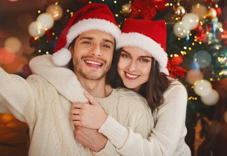Sweet married couple taking selfie on Christmas eve, wearing santa hats over Christmas treeの写真素材