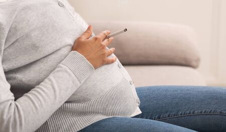 Smoking In Pregnancy. Unrecognizable Pregnant Lady Holding Cigarette Sitting On Sofa At Home. Croppedの写真素材