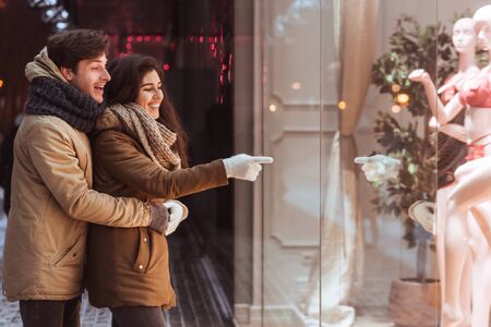 Loving Couple Doing Winter Shopping Standing Outdoor Of A Store And Pointing Finger At Shopwindow In Evening. Empty Spaceの写真素材