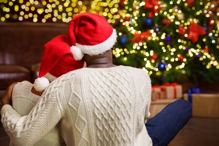 Christmas eve. Rear view of african american couple in Santa hats sitting on floor and looking at Xmas tree, waiting for miraclesの写真素材