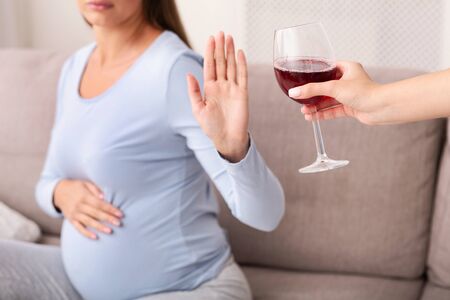 No Alcohol During Pregnancy. Unrecognizable Pregnant Woman Gestring Stop To Glass Of Wine Sitting On Sofa Indoor. Croppedの写真素材